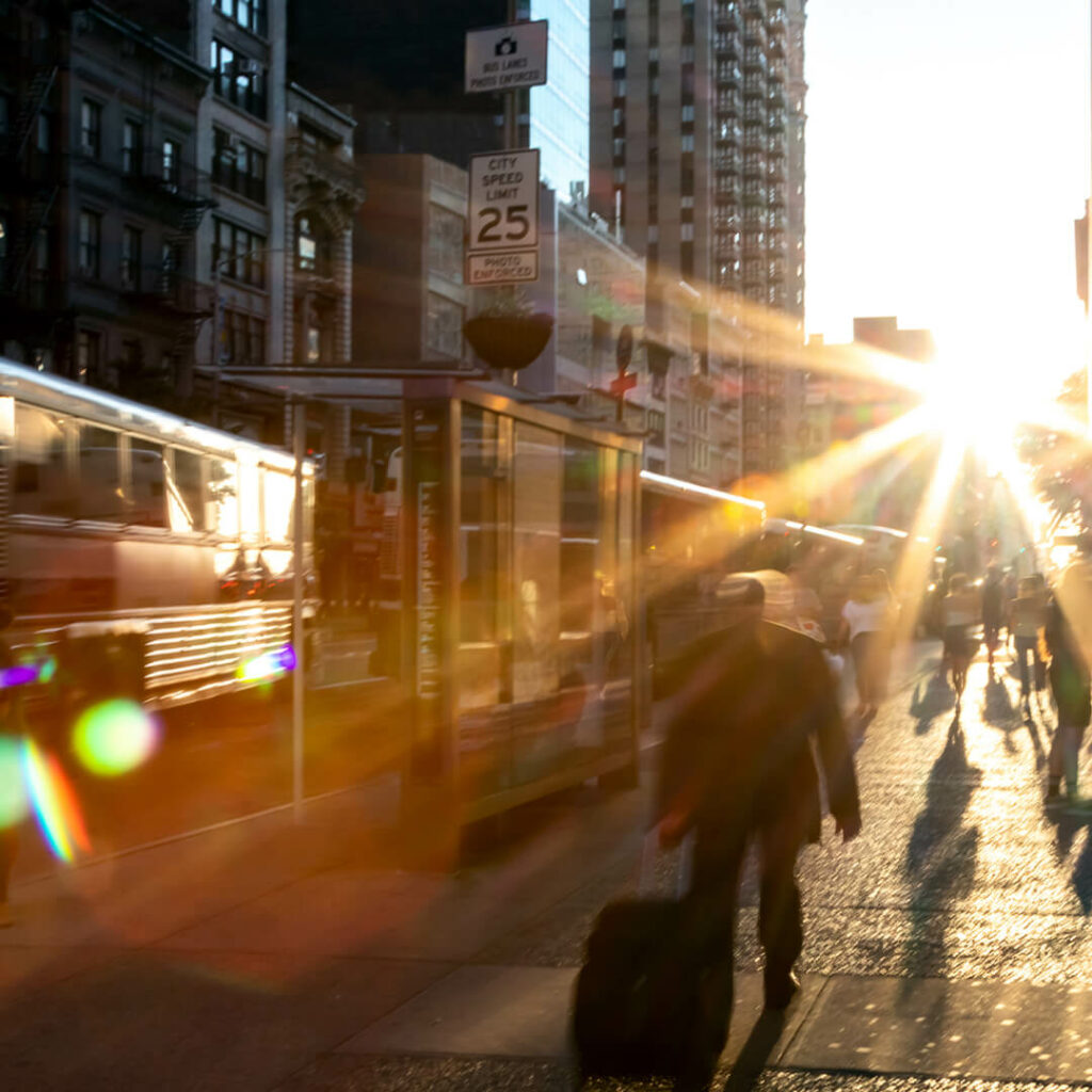 people walking beside a new york street at sunset with buses passing by